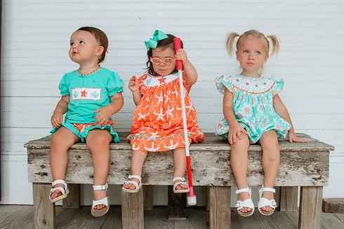 Three toddler girls sitting on bench wearing coral & teal summer smocked outfits.