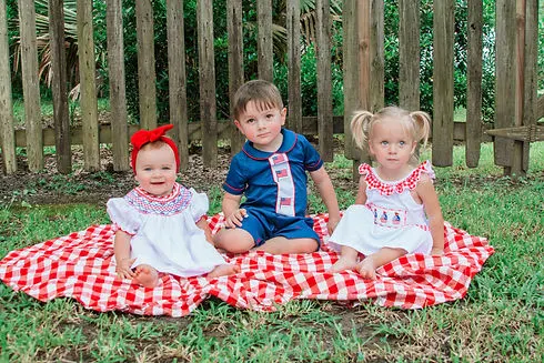 Baby girl, toddler boy, & toddler girl sit on blanket in grass wearing smocked American outfits